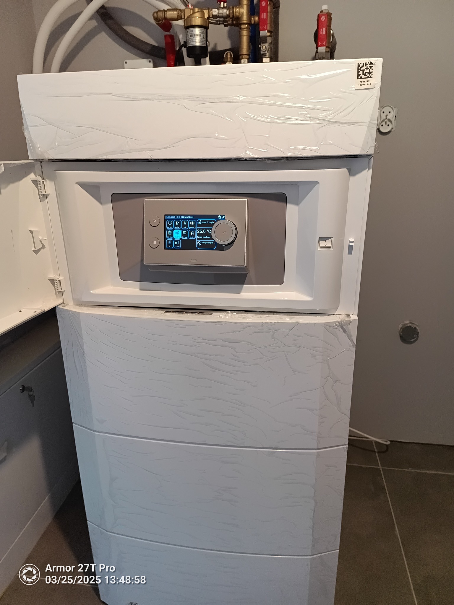 Nowoczesny, biały piec grzewczy z otwartymi drzwiczkami, ukazujący panel sterowania z wyświetlaczem. Widoczne rury instalacji nad urządzeniem.