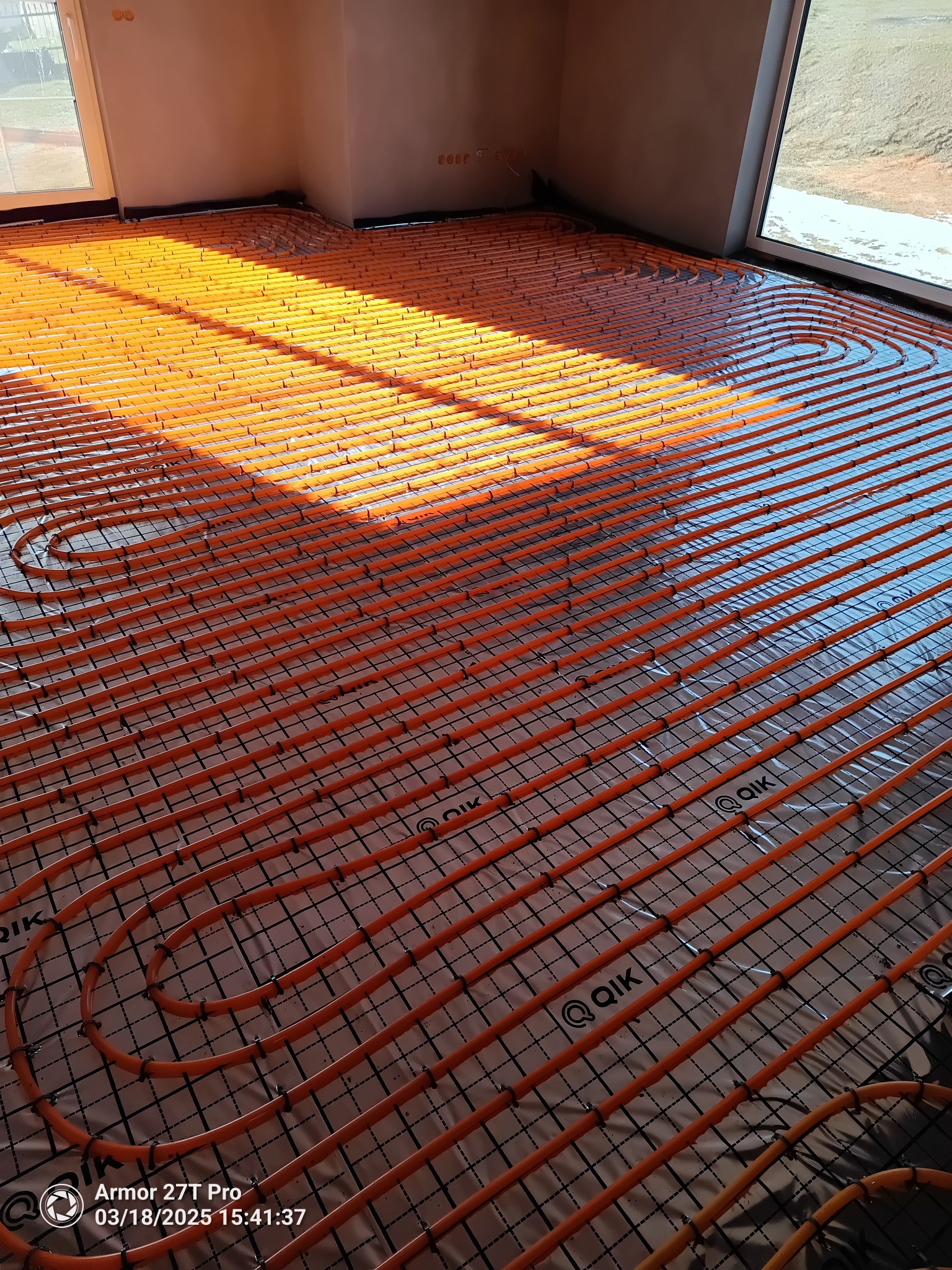 Pomarańczowe rury ogrzewania podłogowego ułożone na folii z siatką, widoczne promienie słoneczne wpadające przez okno,  wnętrze w trakcie wykończenia.