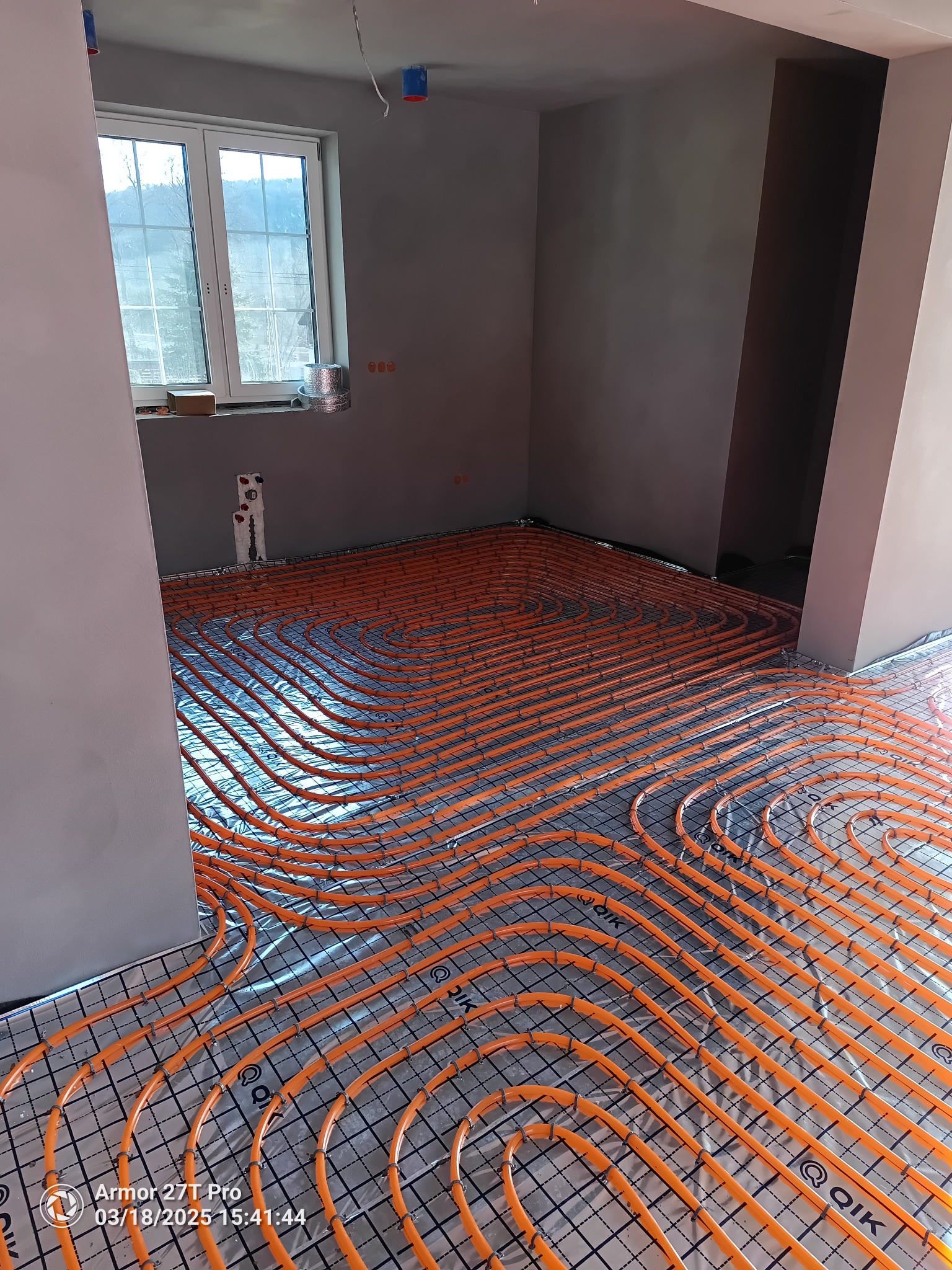 Pomarańczowe rury ogrzewania podłogowego ułożone na srebrnej folii w surowym wnętrzu. Widoczne okno i szare ściany, instalacja w trakcie realizacji.