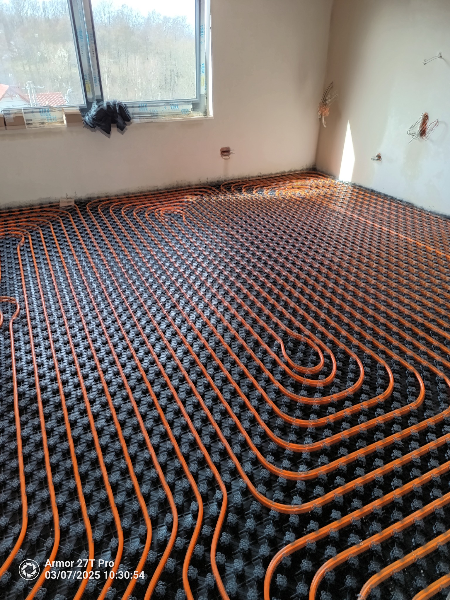 Pomarańczowe rury ogrzewania podłogowego ułożone na czarnej macie w jasnym pomieszczeniu z oknem. Widok z góry ukazuje regularny wzór instalacji.