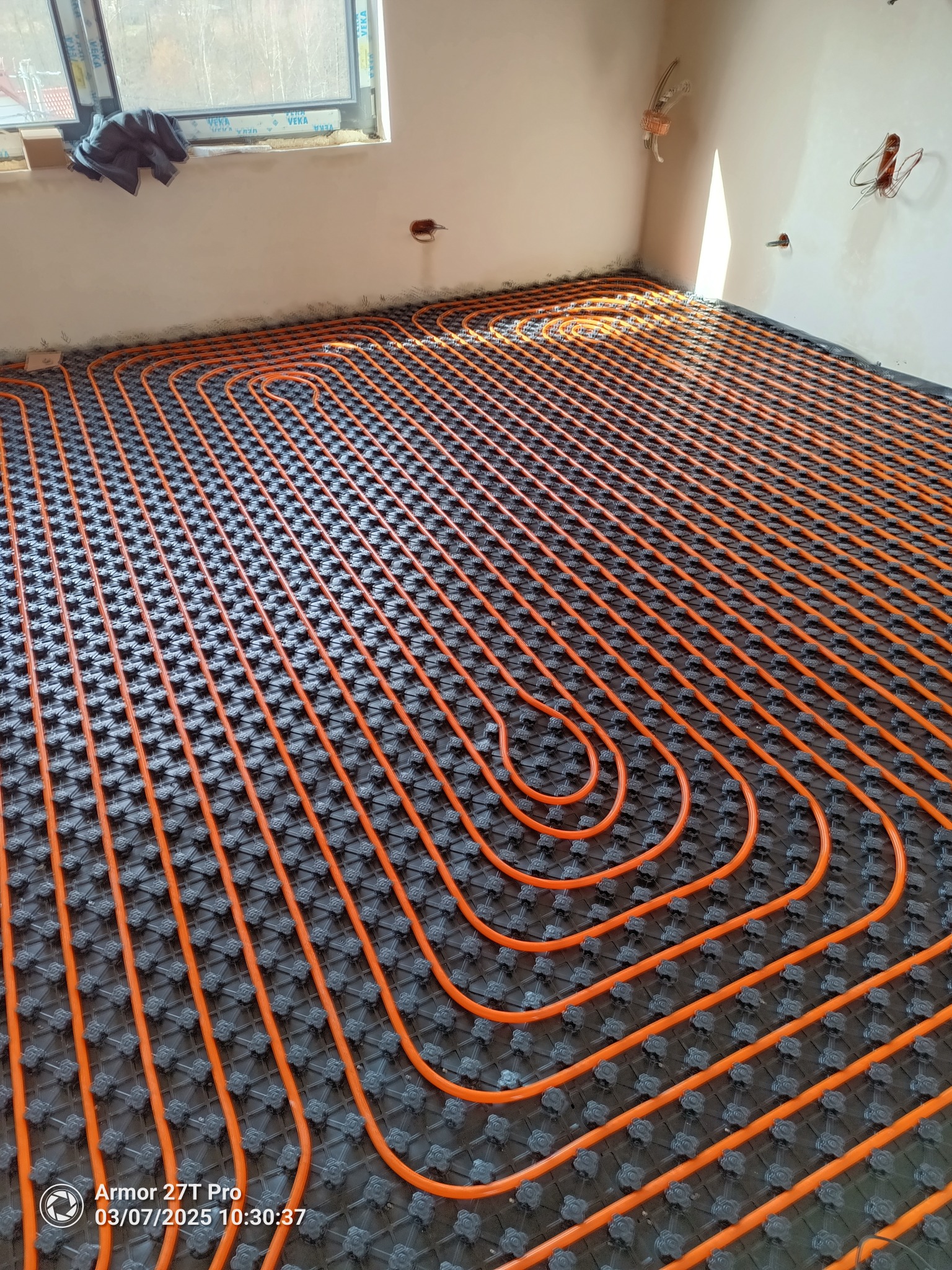 Pomarańczowe rury ogrzewania podłogowego ułożone na czarnej macie w nowym budynku, widoczne kable elektryczne wystające ze ściany, okno w tle.