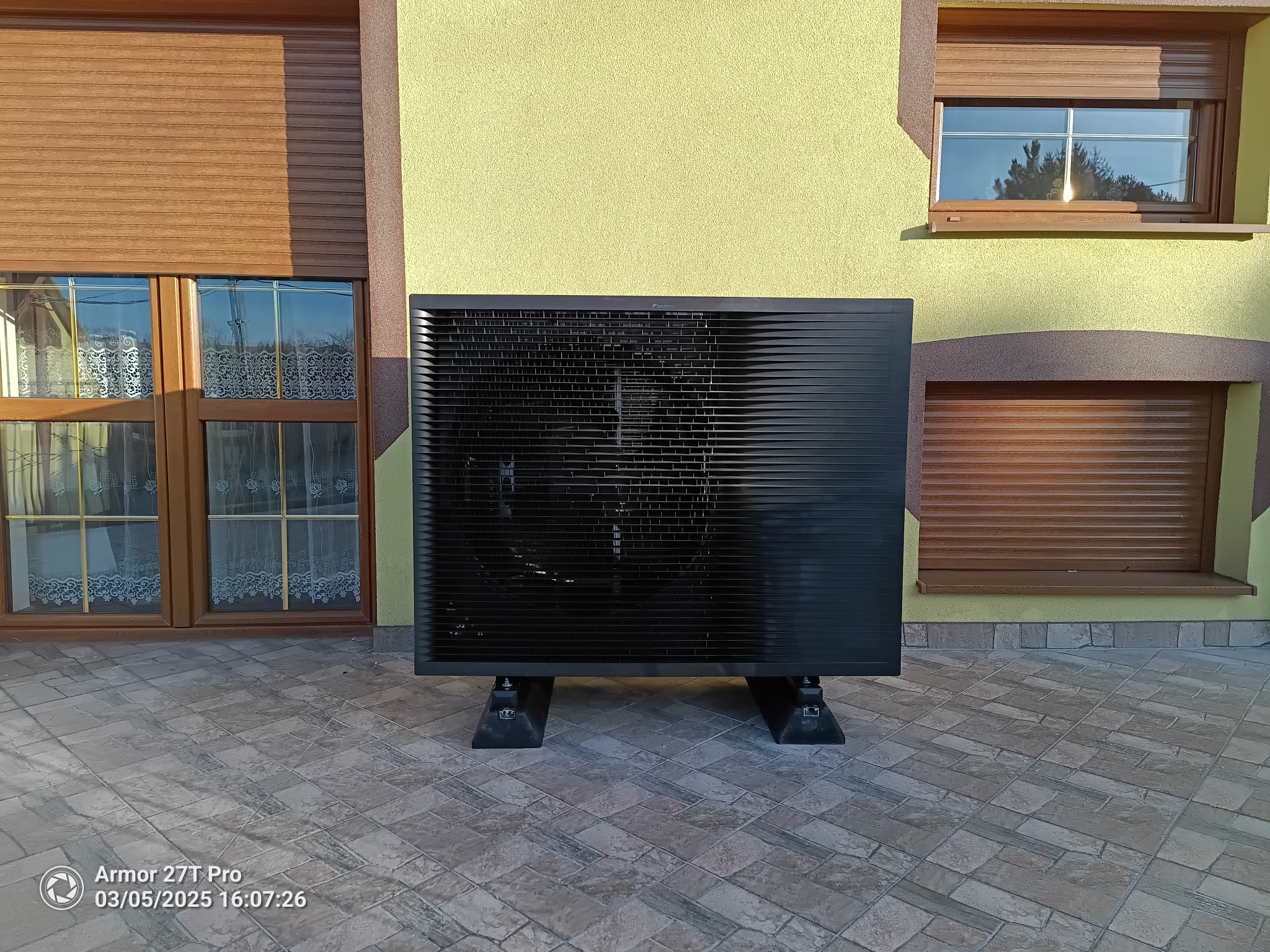 Czarna pompa ciepła Daikin Altherma 3 R ECH2O, zamontowana na zewnątrz domu z żółtą elewacją i brązowymi okiennicami, na tle kostki brukowej.