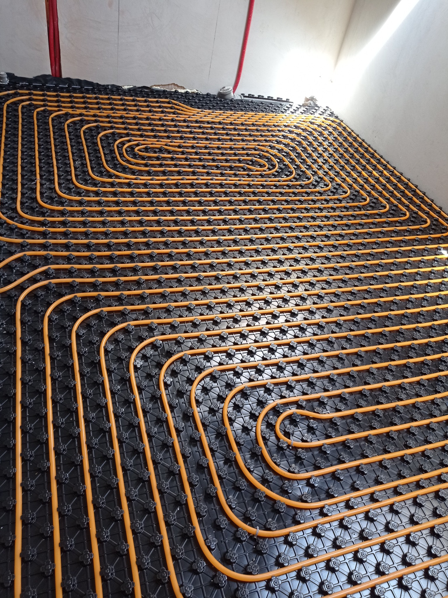 Żółte rury ogrzewania podłogowego ułożone spiralnie na czarnej macie montażowej, gotowe do zalania wylewką. Widok z góry, bliski kadr.