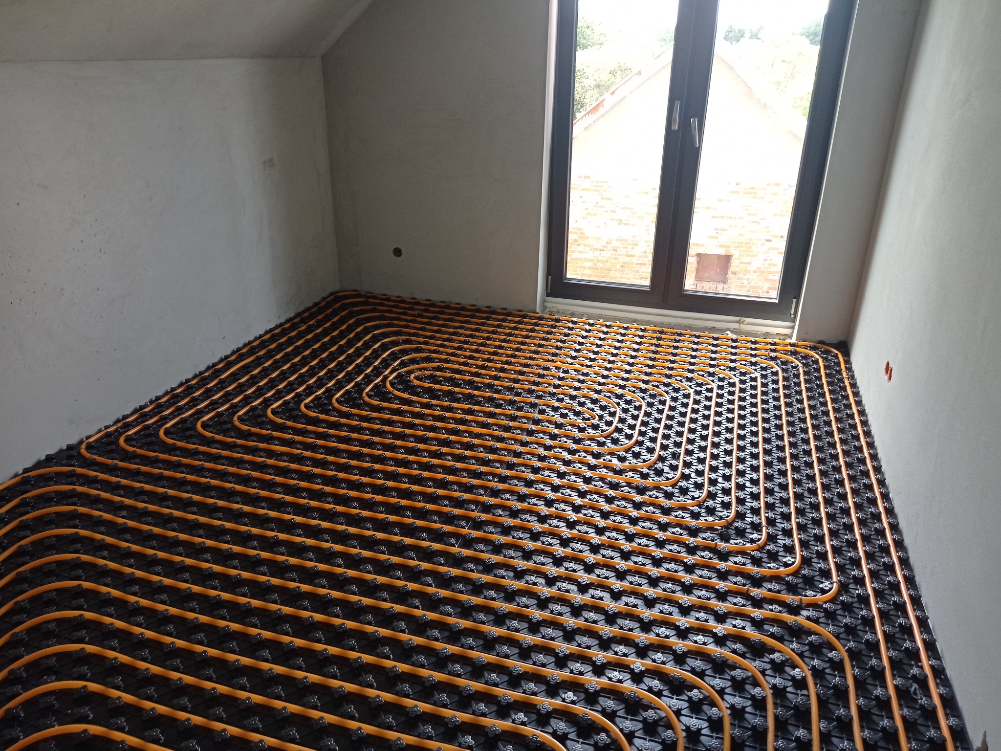 Pomarańczowe rury ogrzewania podłogowego ułożone spiralnie na czarnej macie w surowym pomieszczeniu z oknem. Widok z góry, blisko ściany.