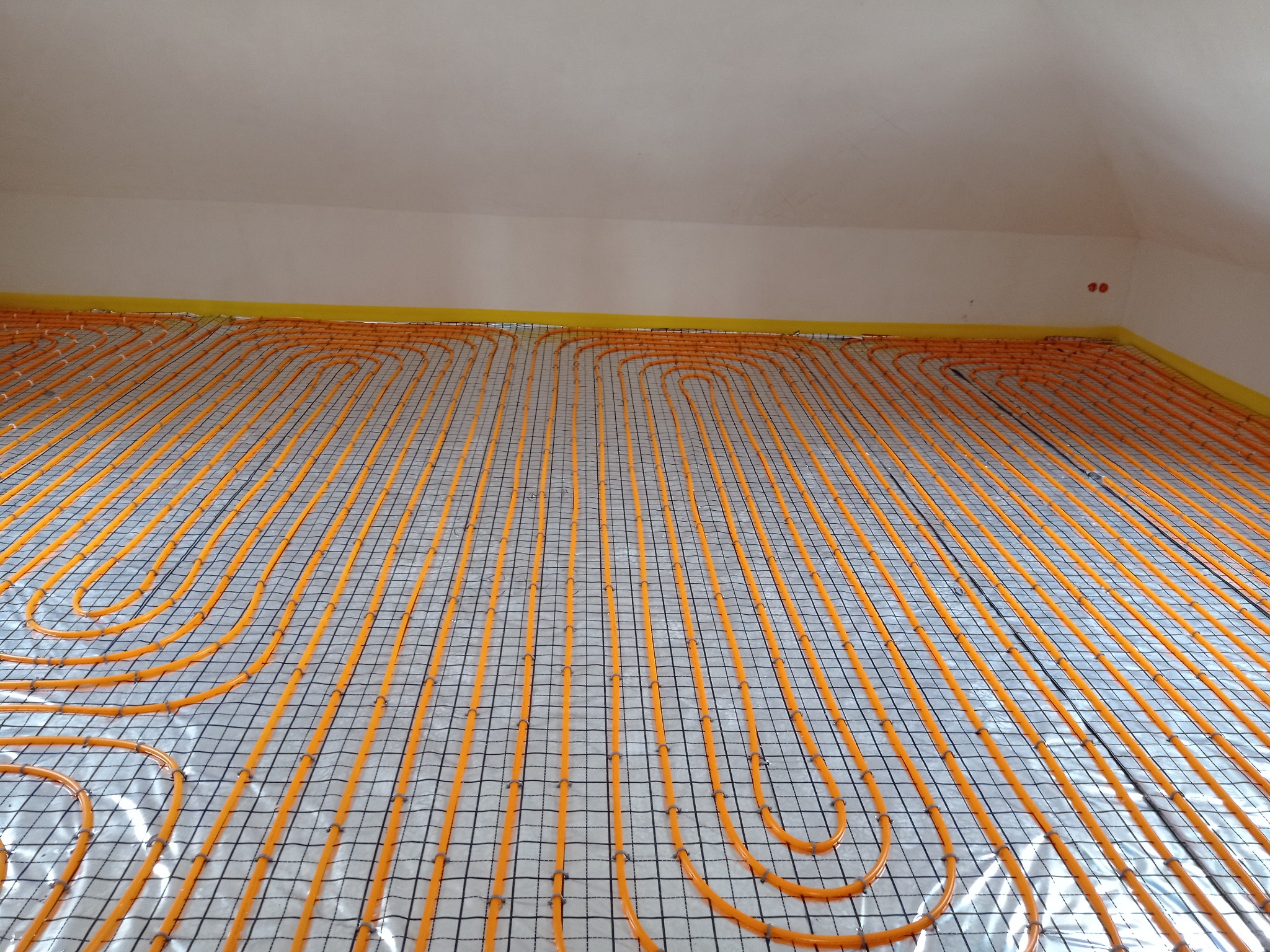 Pomarańczowe rury ogrzewania podłogowego ułożone spiralnie na siatce, gotowe do zalania wylewką. Żółta taśma przy ścianach. Widok z góry, ukośnie.