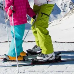 Dwie osoby na nartach na tle ośnieżonych gór, widoczne narty, buty narciarskie i kije, jedna osoba w różowej kurtce i turkusowych spodniach, druga w białej kurtce i limonkowych spodniach.