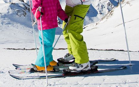 Dwie osoby na nartach na tle ośnieżonych gór, widoczne narty, buty narciarskie i kije, jedna osoba w różowej kurtce i turkusowych spodniach, druga w białej kurtce i limonkowych spodniach.