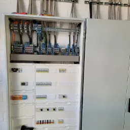 Otwarta szafa rozdzielcza z widocznymi bezpiecznikami, przewodami i elementami sterującymi, zamontowana na ścianie z cegły, z narzędziami na górze.