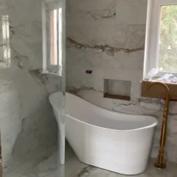 Wanna wolnostojąca w łazience wykończonej marmurowymi płytkami, złota bateria przy oknie, częściowo przeszklona kabina prysznicowa.