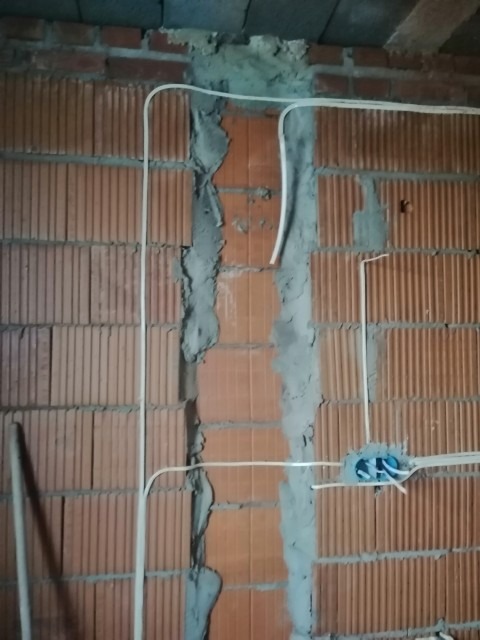 Wewnętrzna instalacja elektryczna w trakcie montażu w bruzdach ceglanej ściany, widoczne przewody w peszlu i puszka elektryczna z niebieskimi złączkami.