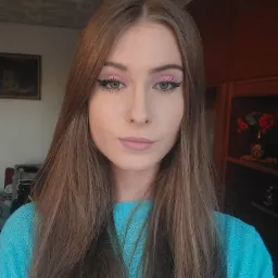 Portret kobiety z długimi brązowymi włosami, w błękitnym swetrze, z różowym makijażem oczu i czarnym eyelinerem, na tle pokoju z obrazem i ciemnymi meblami.