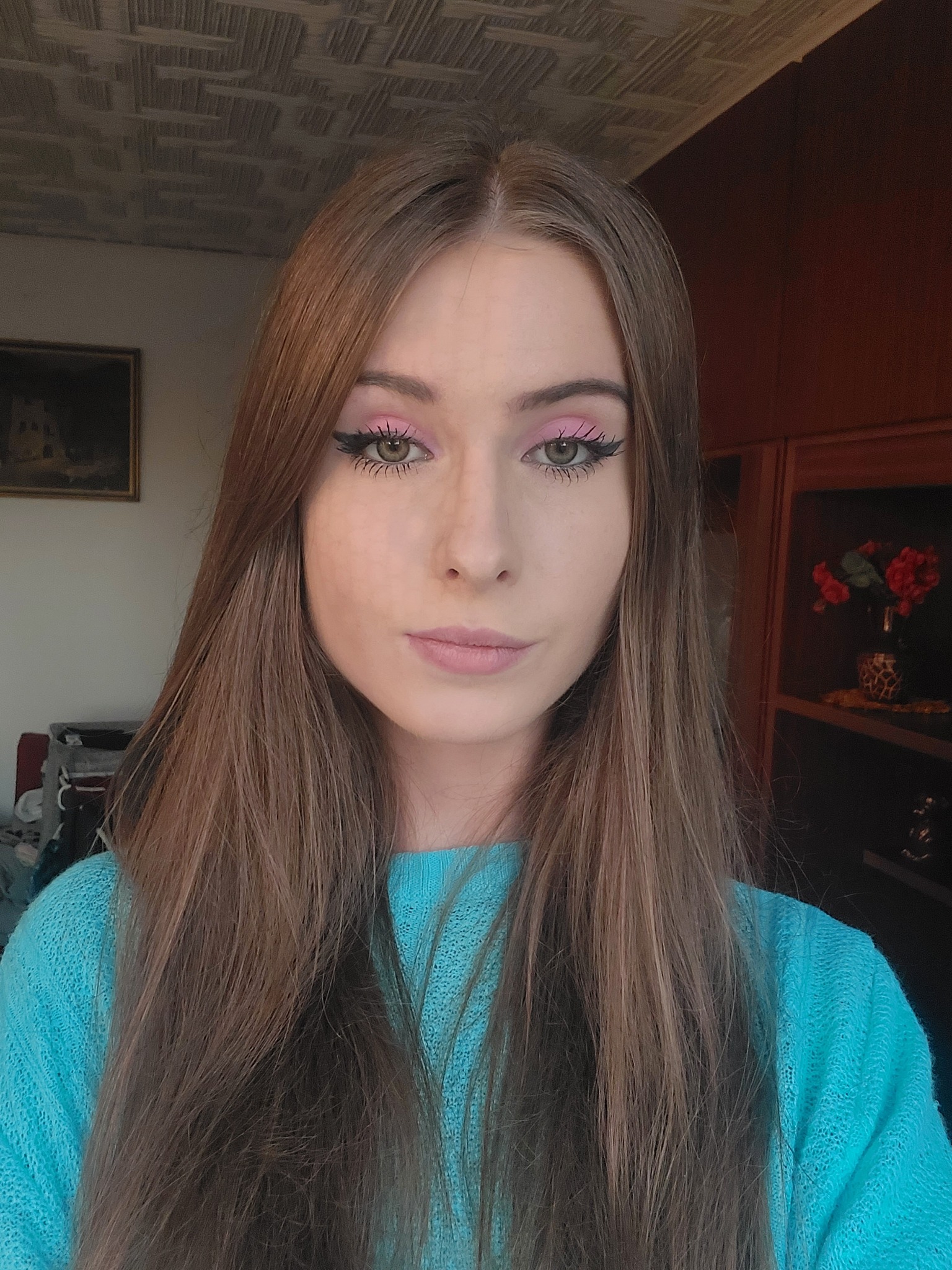 Portret kobiety z długimi brązowymi włosami, w błękitnym swetrze, z różowym makijażem oczu i czarnym eyelinerem, na tle pokoju z obrazem i ciemnymi meblami.