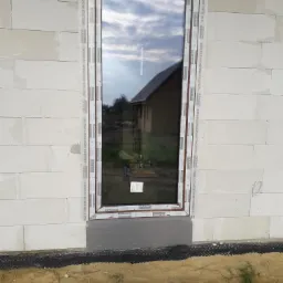 Montaż okna w Siedlcach w surowym murze z betonu komórkowego; widoczne folie montażowe na ramie okiennej i izolacja fundamentu.