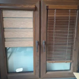 Brązowe okno z dwiema różnymi osłonami: roletą dzień noc i żaluzjami aluminiowymi, widoczne na tle jasnych płytek podłogowych, z naklejką na szybie.