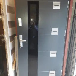 Szare drzwi wejściowe z długim, pionowym przeszkleniem i czterema kwadratowymi, metalowymi elementami dekoracyjnymi, widoczny zamek z klamką i kluczami, odbicie fotografa w szybie.