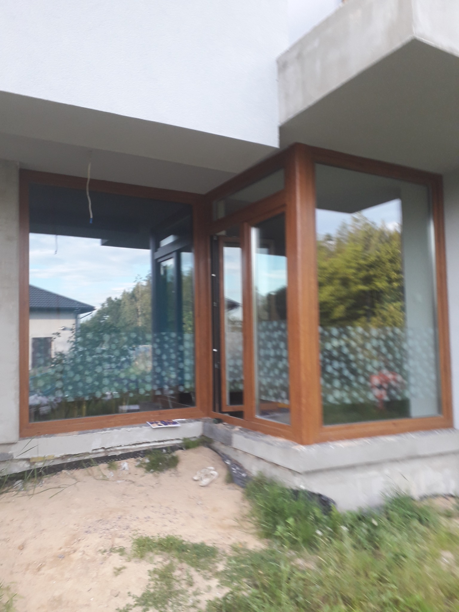 Narożne okno z brązowymi ramami w surowym stanie, widoczne folie ochronne na szybach i piasek przed budynkiem.