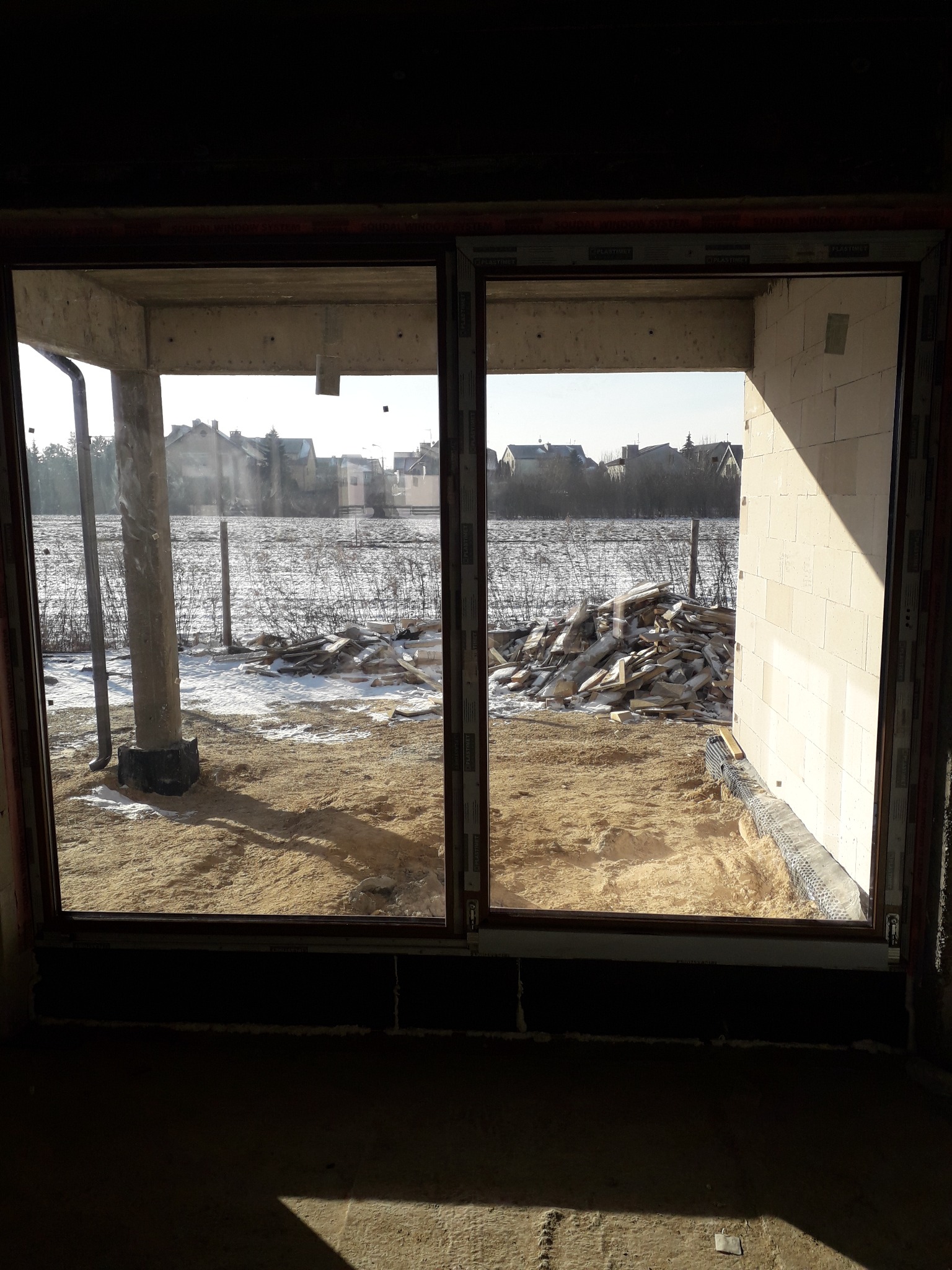 Widok przez dwuskrzydłowe okno na plac budowy w zimowej scenerii, z widocznymi elementami konstrukcyjnymi i stertą materiałów budowlanych na zewnątrz.