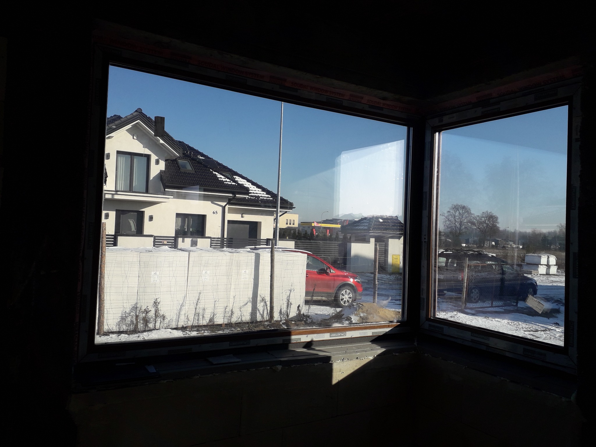 Widok z wnętrza budynku w trakcie budowy przez nowo zamontowane okna na dom z garażem, zaparkowane samochody i materiały budowlane.