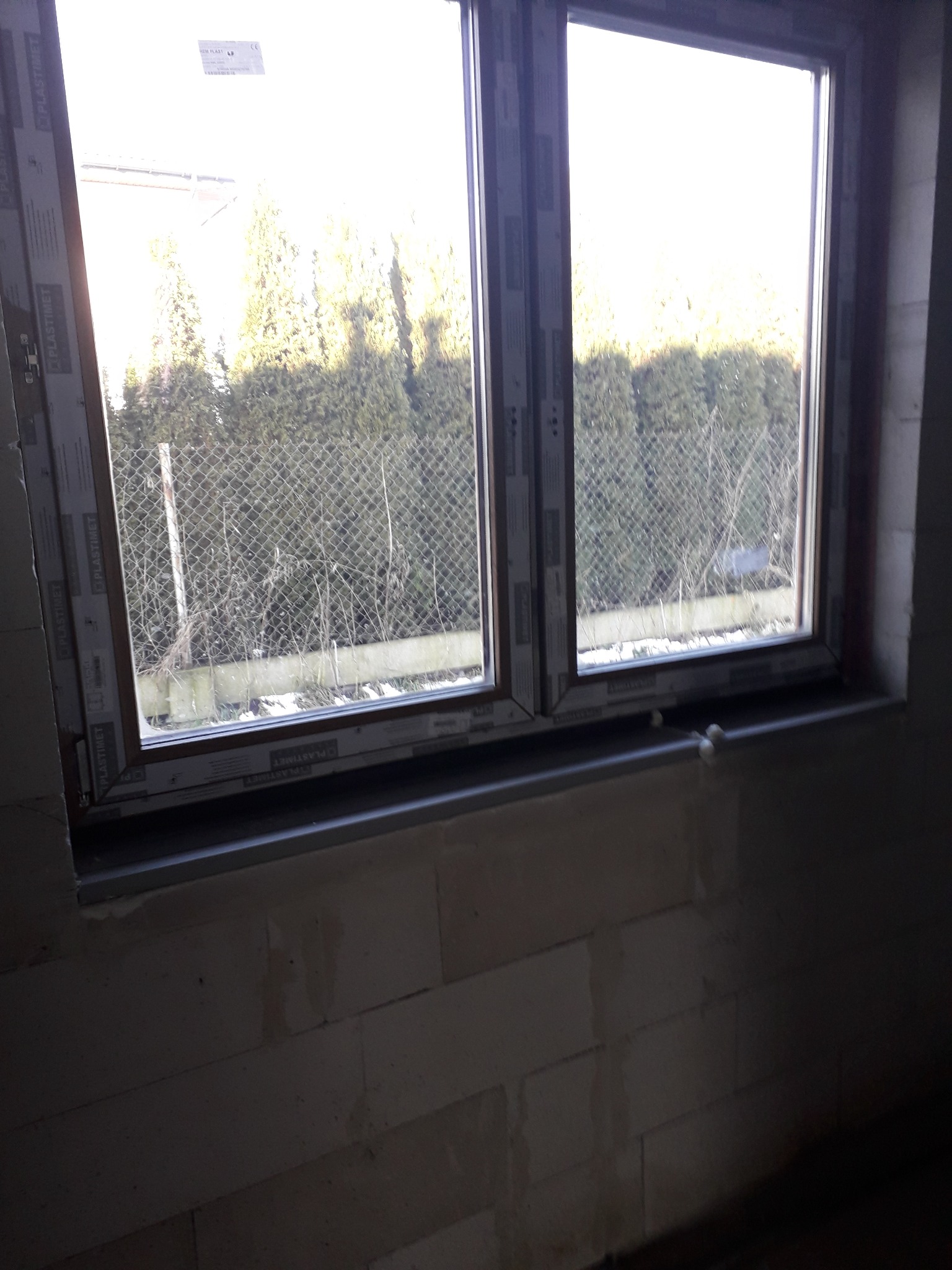 Montaż dwuskrzydłowego okna z brązową ramą, widok na zewnątrz z siatką ogrodzeniową i drzewami w tle, ściany z bloków betonu komórkowego w trakcie wykończenia.