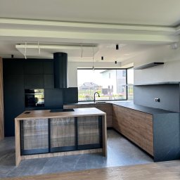 OLIWIA GUMIŃSKA OLIV. DESIGN - Nowoczesna kuchnia z wyspą: czarne szafki, drewnopodobne fronty, szklane witryny w wyspie, czarny okrągły okap, duże okno z widokiem na ogród.