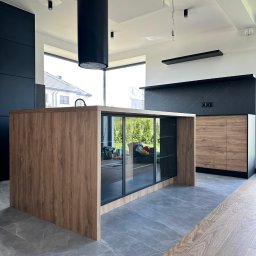 OLIWIA GUMIŃSKA OLIV. DESIGN - Nowoczesna kuchnia z wyspą o drewnianym wykończeniu i przeszklonymi szafkami, z widocznym wyposażeniem wnętrza i czarnym okapem cylindrycznym.