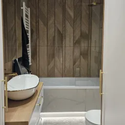 Nowoczesna łazienka z drewnopodobnymi płytkami ułożonymi w jodełkę na ścianie, białą umywalką nablatową z złotymi akcentami, podświetlaną wanną i miękkim dywanikiem.