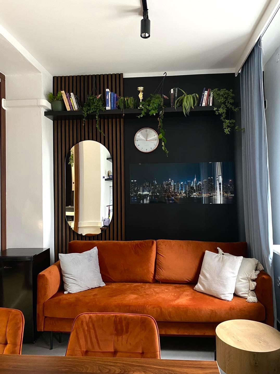 Salon z pomarańczową sofą, lustrem w drewnianej ramie, czarną ścianą z półką, roślinami, zegarem i panoramą miasta nocą.