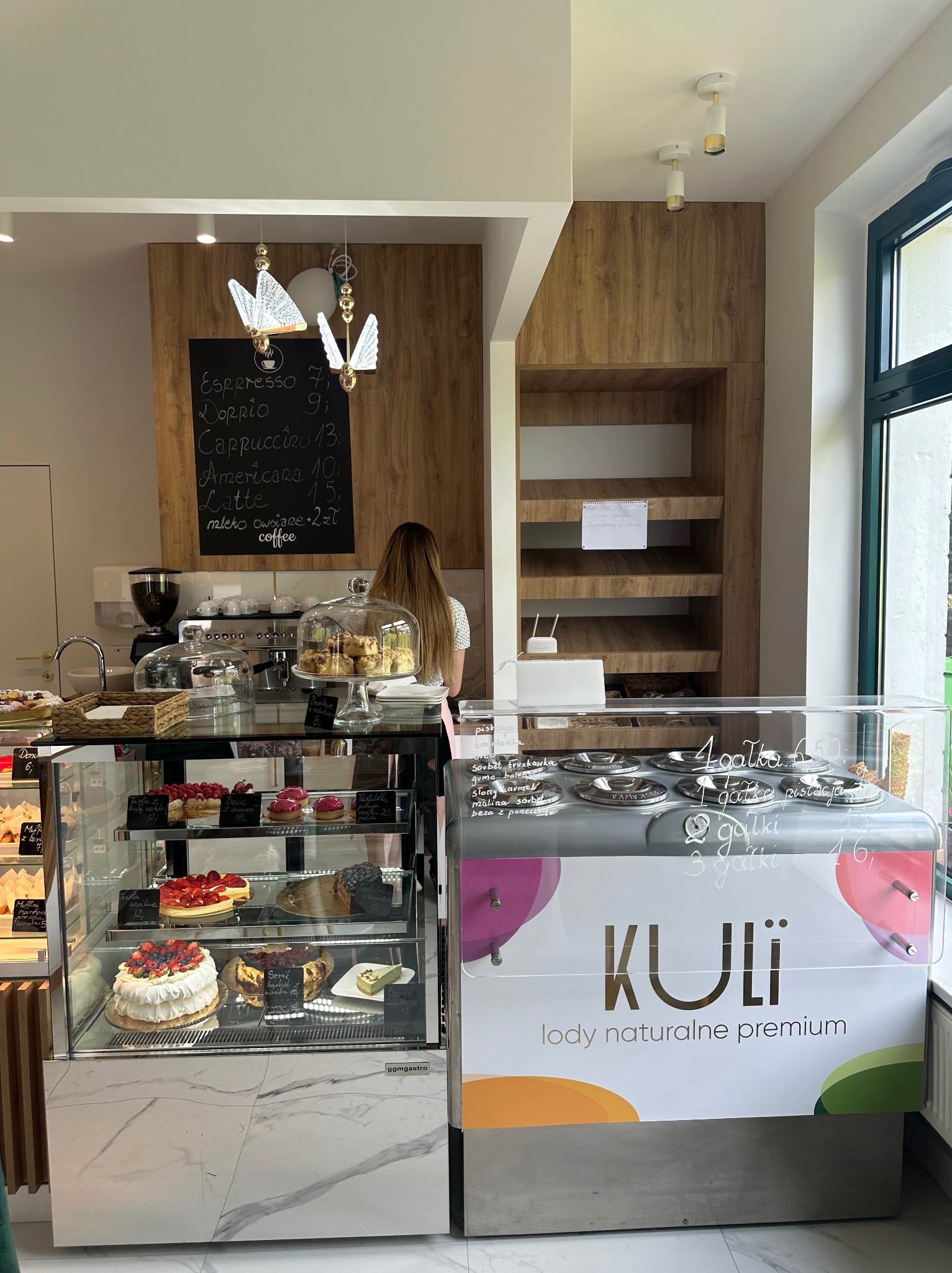 Wnętrze kawiarni z ekspozycją ciast i lodów, menu wypisane kredą na tablicy, oświetlenie w kształcie motyli, nowoczesny design.