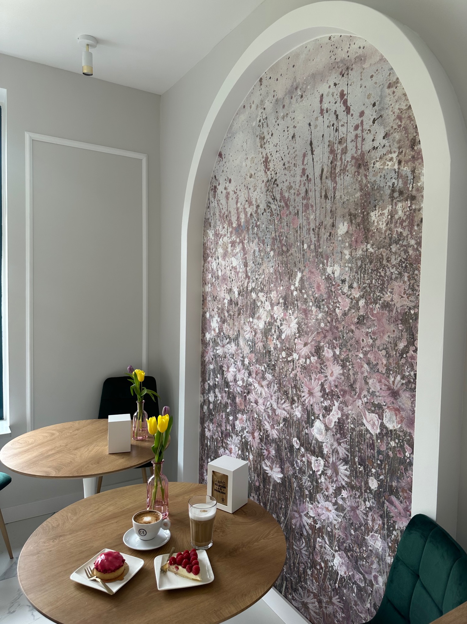 Jasne wnętrze kawiarni z okrągłymi stolikami, deserami i kawą, ozdobione tapetą imitującą łąkę kwiatów za łukiem w ścianie.