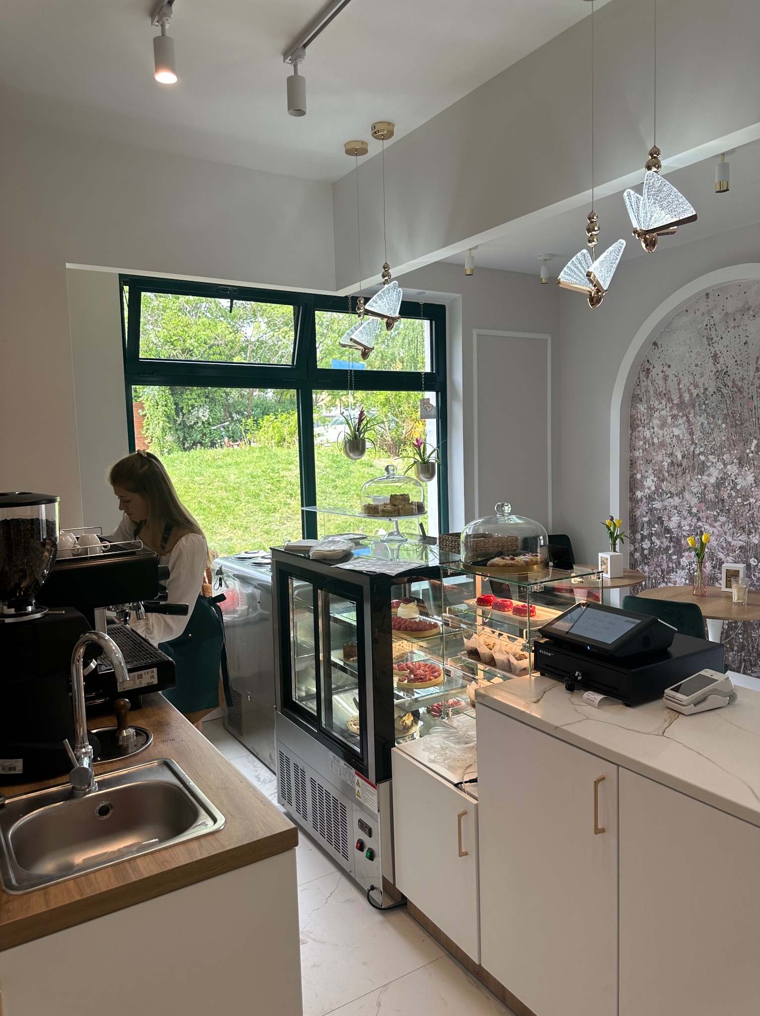 Jasne wnętrze kawiarni z ekspozycją ciast i deserów w chłodzonej witrynie, za ladą barista przygotowuje kawę, okno z widokiem na zieleń.