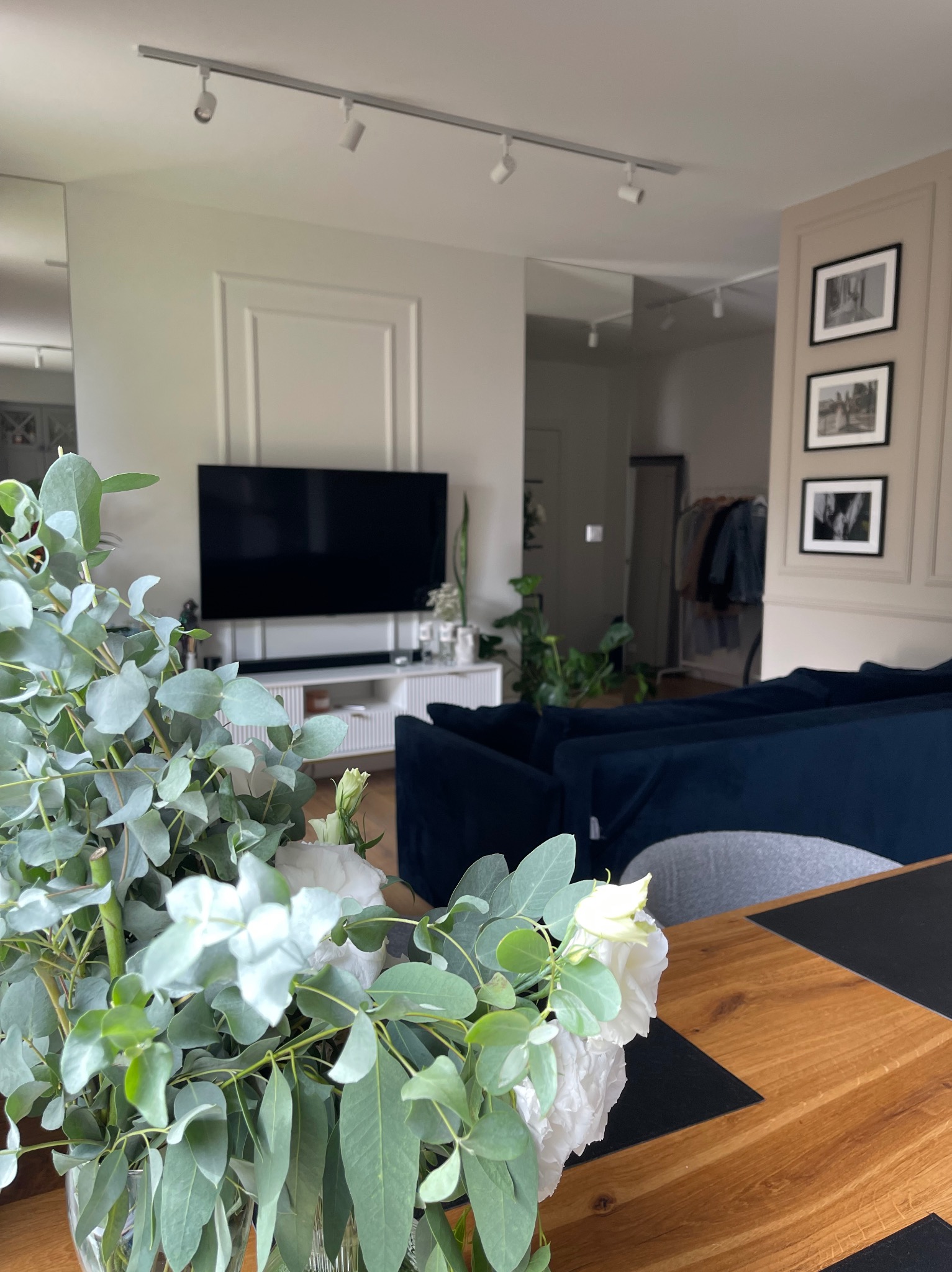 Salon z ciemnoniebieską sofą, telewizorem na białej szafce RTV, oświetleniem szynowym i dekoracyjnymi panelami ściennymi; na pierwszym planie bukiet eukaliptusa i białych kwiatów na drewnianym stole.