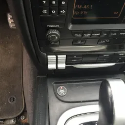 Wnętrze samochodu marki Porsche z widocznym panelem sterowania, dźwignią zmiany biegów i fragmentem podłogi z widocznymi elementami mocowania. Panel radia wyświetla napis 'OSTSEE'.