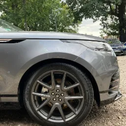 Szary samochód Range Rover Velar z aluminiowymi felgami na żwirowym podłożu, widok z boku na przednie koło i fragment nadwozia.
