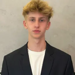 Portret młodego mężczyzny w czarnej marynarce i białej koszulce, o blond włosach zaczesanych do góry, na tle jasnej ściany.