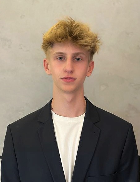 Portret młodego mężczyzny w czarnej marynarce i białej koszulce, o blond włosach zaczesanych do góry, na tle jasnej ściany.