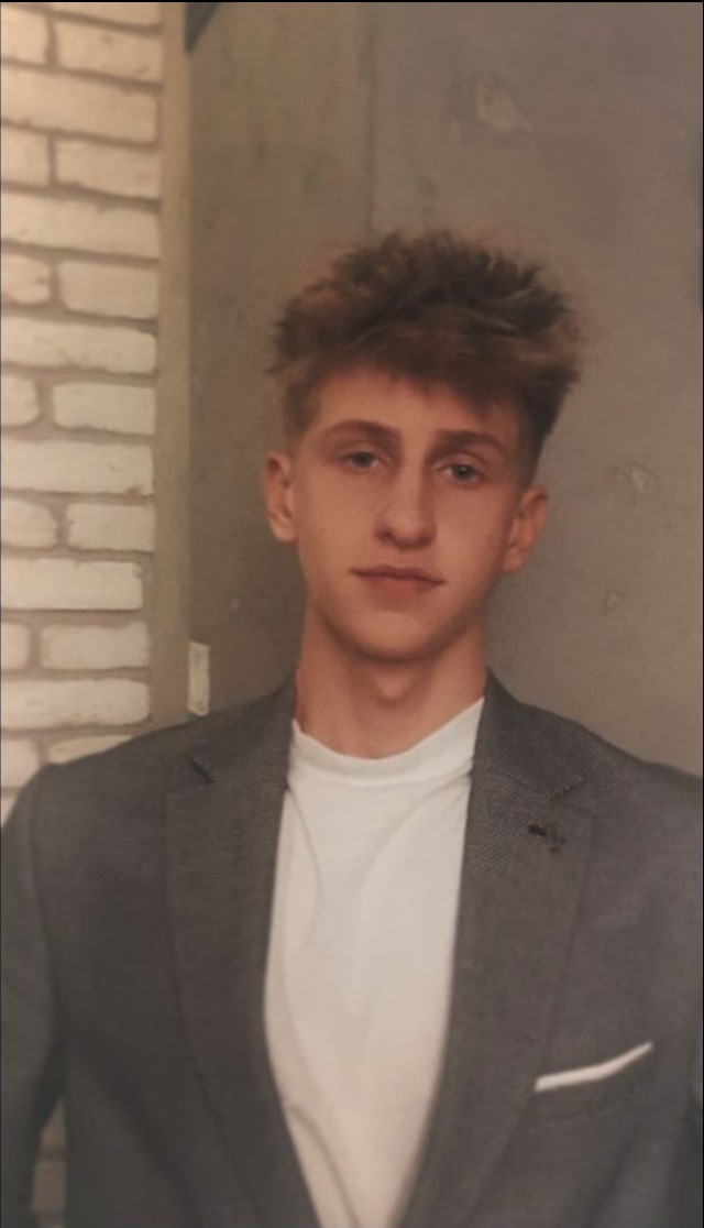 Młody mężczyzna w szarej marynarce i białym t-shircie pozuje na tle ceglanej ściany.