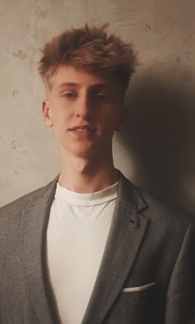 Portret młodego mężczyzny z blond włosami, ubranego w szarą marynarkę i biały t-shirt, na tle szarej ściany.