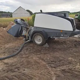 Agregat do wylewania posadzek jastrychowych stoi na błotnistym terenie budowy, z widocznym stosem piasku i budynkiem w tle.