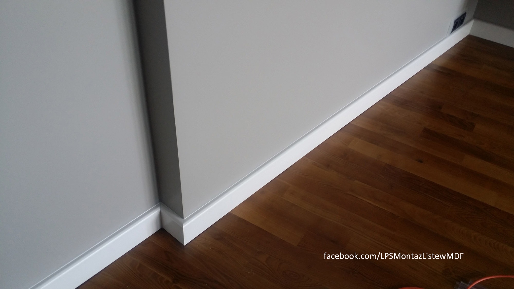 Szary narożnik pokoju z zamontowanymi białymi listwami przypodłogowymi MDF na brązowej podłodze z paneli.