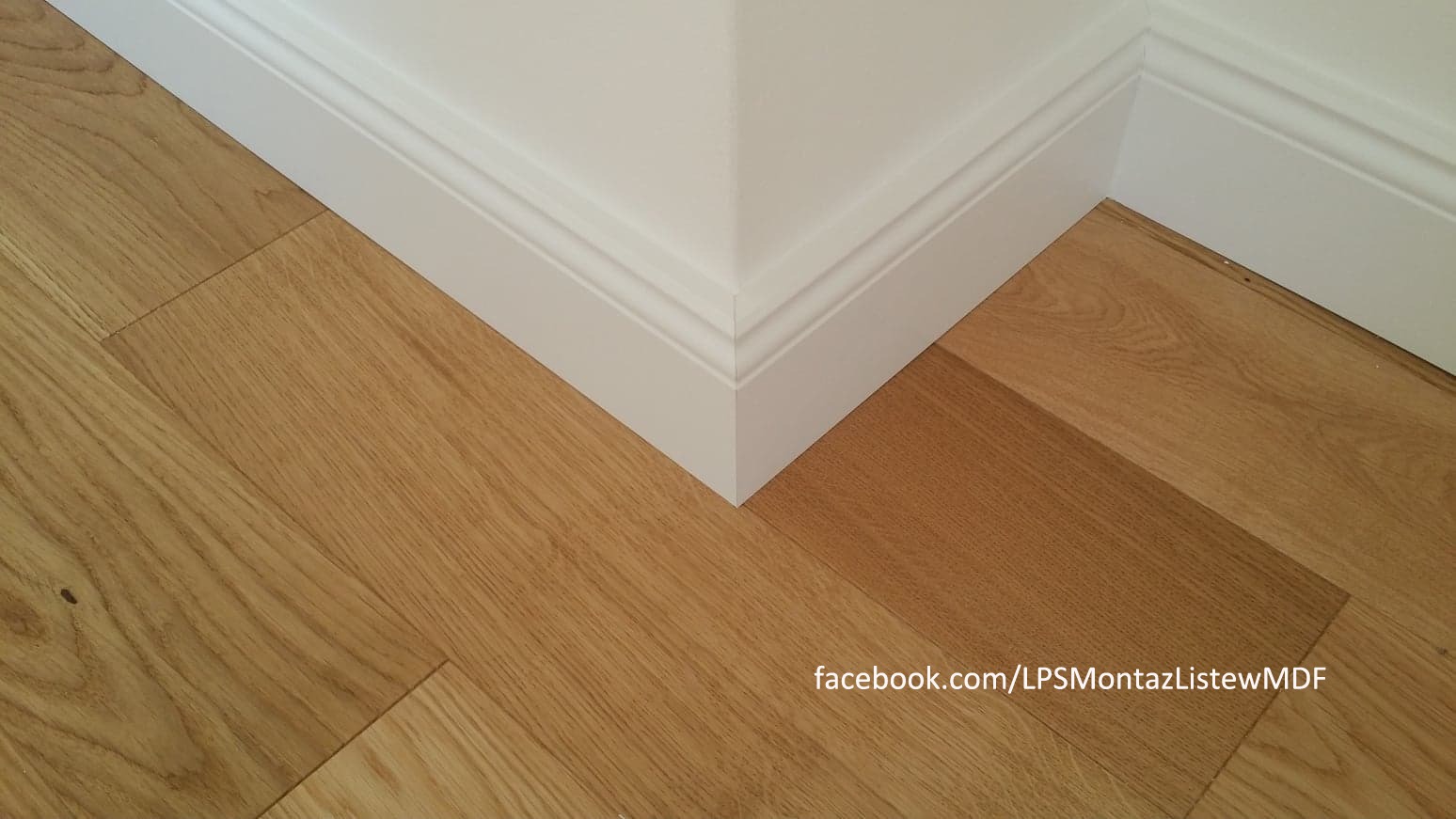 Zamontowane białe listwy przypodłogowe MDF przy drewnianej podłodze w rogu pokoju, widoczne delikatne łączenie paneli podłogowych.