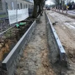 Wykop z betonowymi obrzeżami przygotowany pod budowę chodnika lub drogi, widoczne drzewa i budynek w tle, teren budowy.