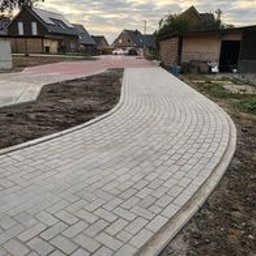 ExpertBruk Brukarstwo Roboty Ziemne - Kremowa ścieżka z kostki brukowej ułożona w łuk, z widocznymi domami w tle i elementami krajobrazu w trakcie prac wykończeniowych.