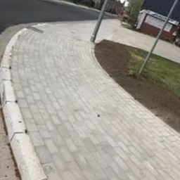 Ułożona jasnoszara kostka brukowa tworząca zakręt chodnika, obrzeża z szarych betonowych elementów, widok z góry na gotową nawierzchnię w otoczeniu trawnika i ulicy.