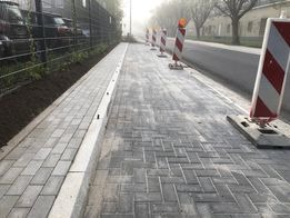 Świeżo ułożona szara kostka brukowa w jodełkę na chodniku, oddzielona białym obrzeżem od trawnika i ulicy, zabezpieczona słupkami ostrzegawczymi, w tle budynki i zaparkowane samochody.