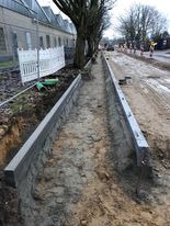 Wykop z betonowymi obrzeżami przygotowany pod budowę chodnika lub drogi, widoczne drzewa i budynek w tle, teren budowy.