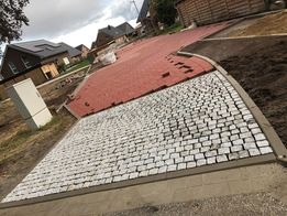 Fragment nowo ułożonej nawierzchni z kostki brukowej w dwóch kolorach: szarej i czerwonej. Szara kostka ułożona w regularny wzór, czerwona w nieregularny. Widoczny obrzeże i przygotowane podłoże.