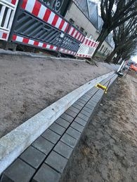 Krawężnik z ułożonym rzędem szarej kostki brukowej wzdłuż wykopu, z widocznymi narzędziami i barierami ochronnymi w tle.