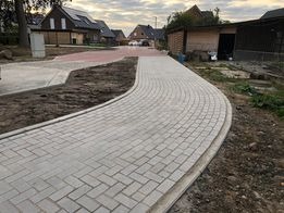 Kremowa ścieżka z kostki brukowej ułożona w łuk, z widocznymi domami w tle i elementami krajobrazu w trakcie prac wykończeniowych.