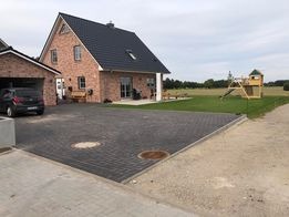 Nowoczesny dom jednorodzinny z ciemną kostką brukową na podjeździe, widoczne studzienki kanalizacyjne, garaż z czerwonej cegły i plac zabaw dla dzieci na trawniku.