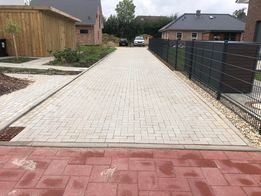 Ułożona, jasnoszara kostka brukowa na długim podjeździe przed domem jednorodzinnym. Czerwona kostka oddziela podjazd od trawnika. Widoczne ogrodzenie panelowe i fragmenty budynków w tle.